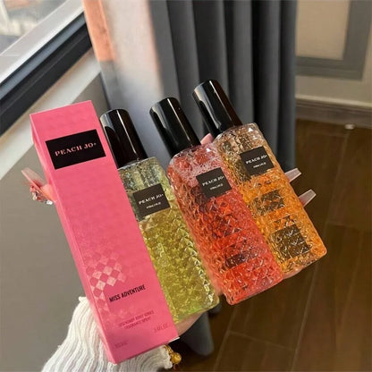 Three perfume bottles with colorful liquids and a pink box labeled 'Peach Joy' on a blurred indoor background.
